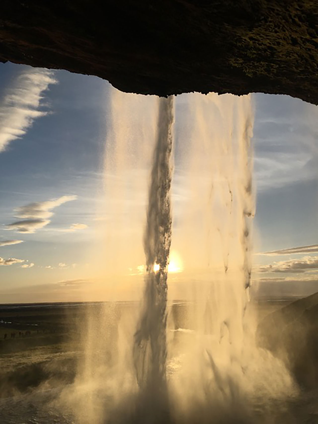 View of a waterfall from behind with sunlight shining through the falling water and a landscape visible in the background.