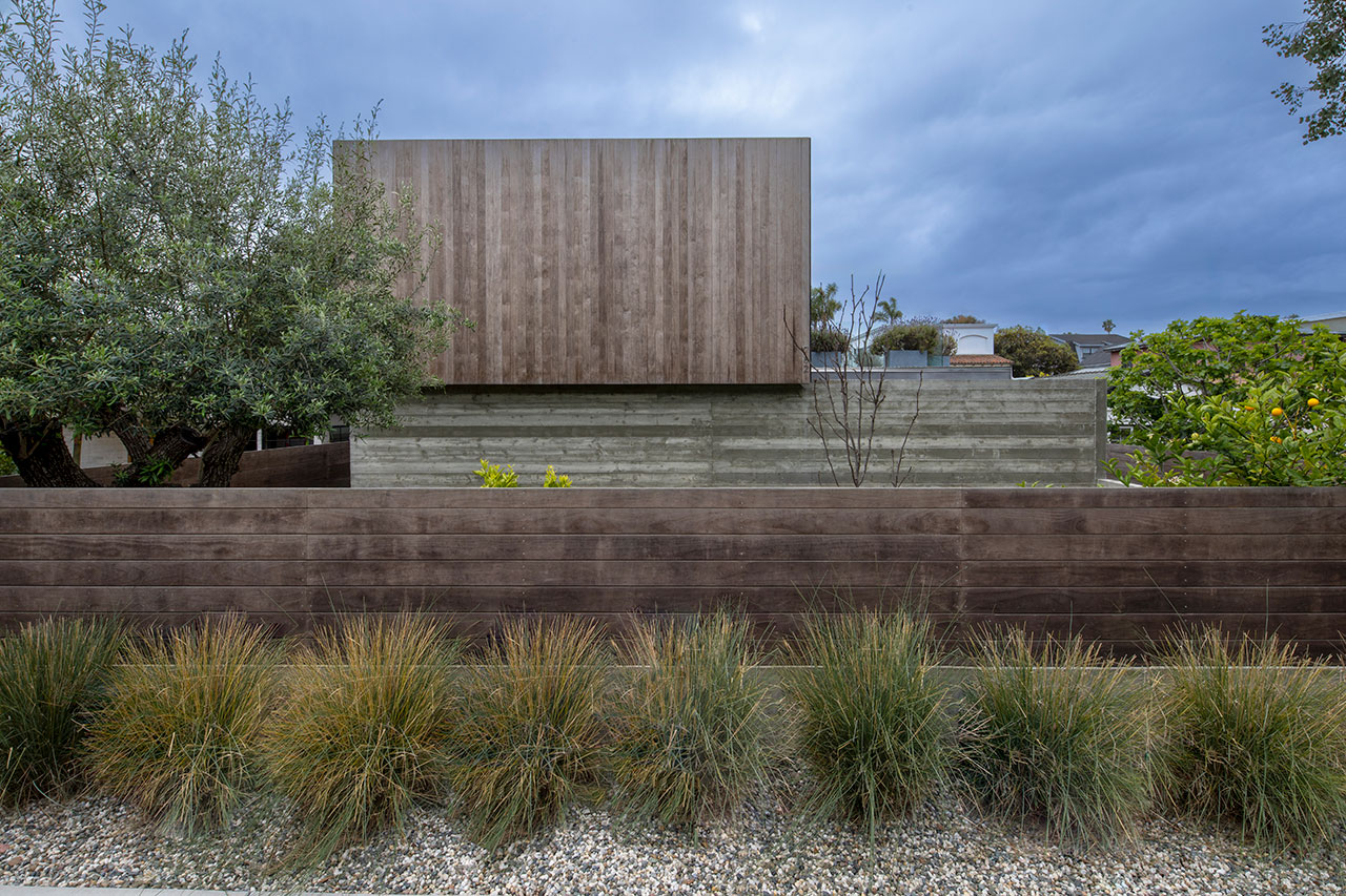 Modern house with a boxy wooden upper section and concrete lower wall, surrounded by a wooden fence, gravel, and ornamental grasses.