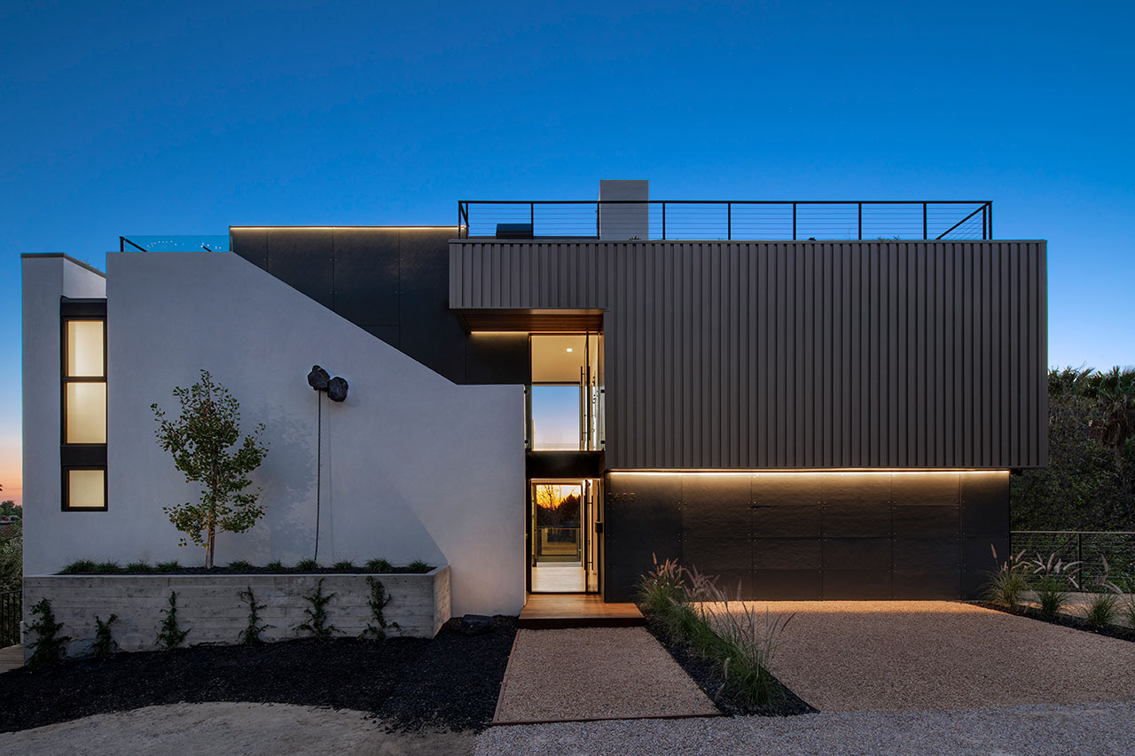 Modern two-story house with flat roof, large windows, and minimalist landscaping, photographed at dusk with illuminated exterior lighting.