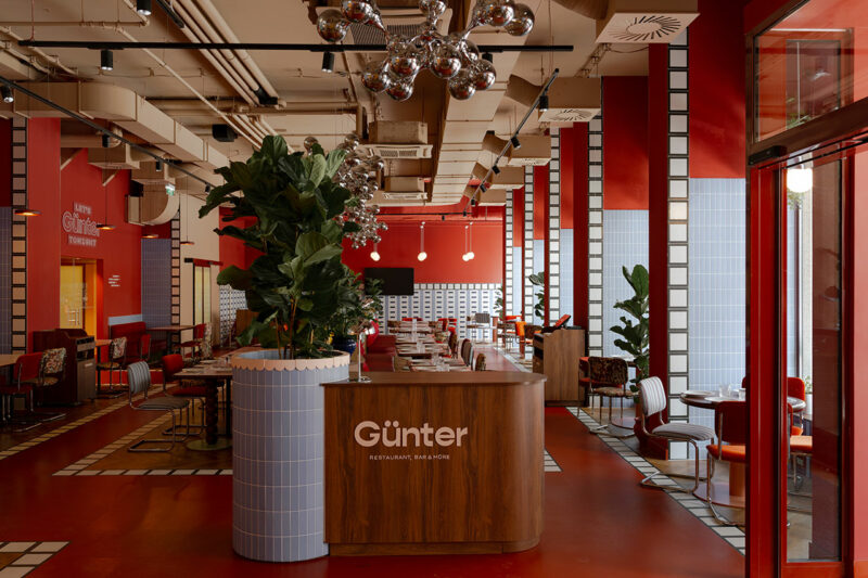 Modern restaurant interior by Atelier OLK with red walls, geometric patterns, large plants, and a wooden reception desk labeled "Günter." Tables and chairs are arranged neatly throughout the space.
