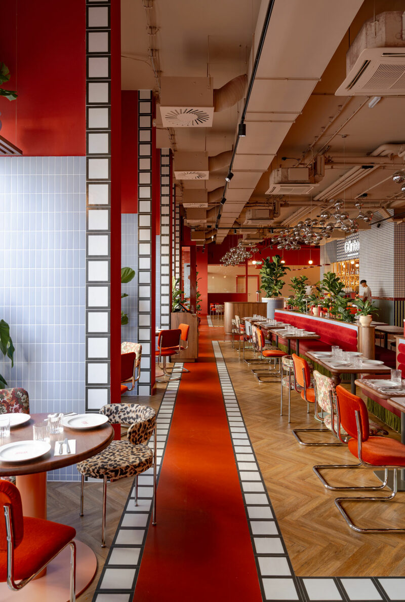 A modern restaurant interior by Atelier OLK, featuring red accents, orange chairs, patterned flooring, white tiled columns, and a long bar lined with stools. Tables are set and plants are placed throughout the stylish space.