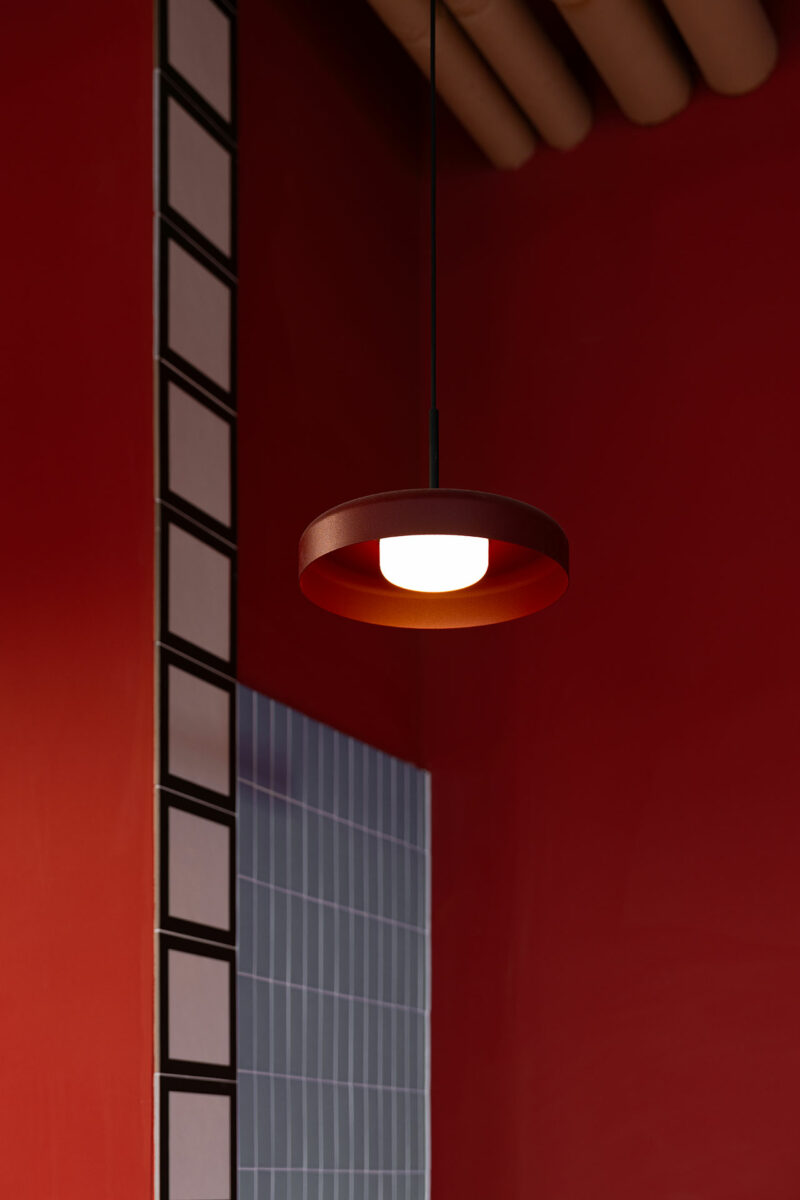 A round, modern pendant light by Atelier OLK hangs from the ceiling in a red room with a tiled wall and a vertical strip of square tiles.