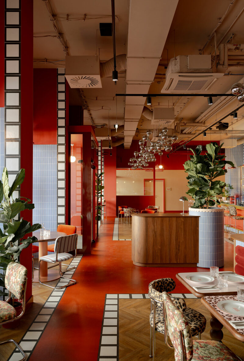 Modern restaurant interior by Atelier OLK with red floors and walls, geometric tile accents, wooden furniture, large potted plants, and patterned chairs arranged around set tables.