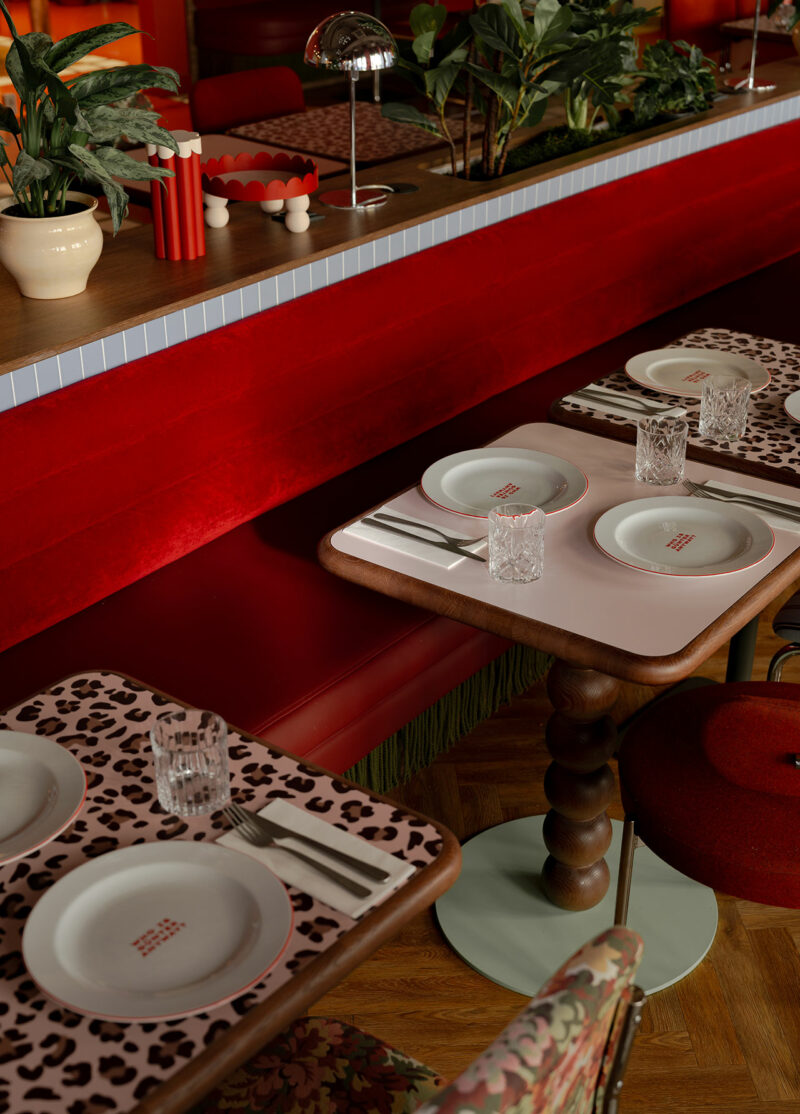 Restaurant table setting with white plates, clear glasses, cutlery, and leopard print placemats by Atelier OLK. Red velvet seating and potted plants are visible in the background.