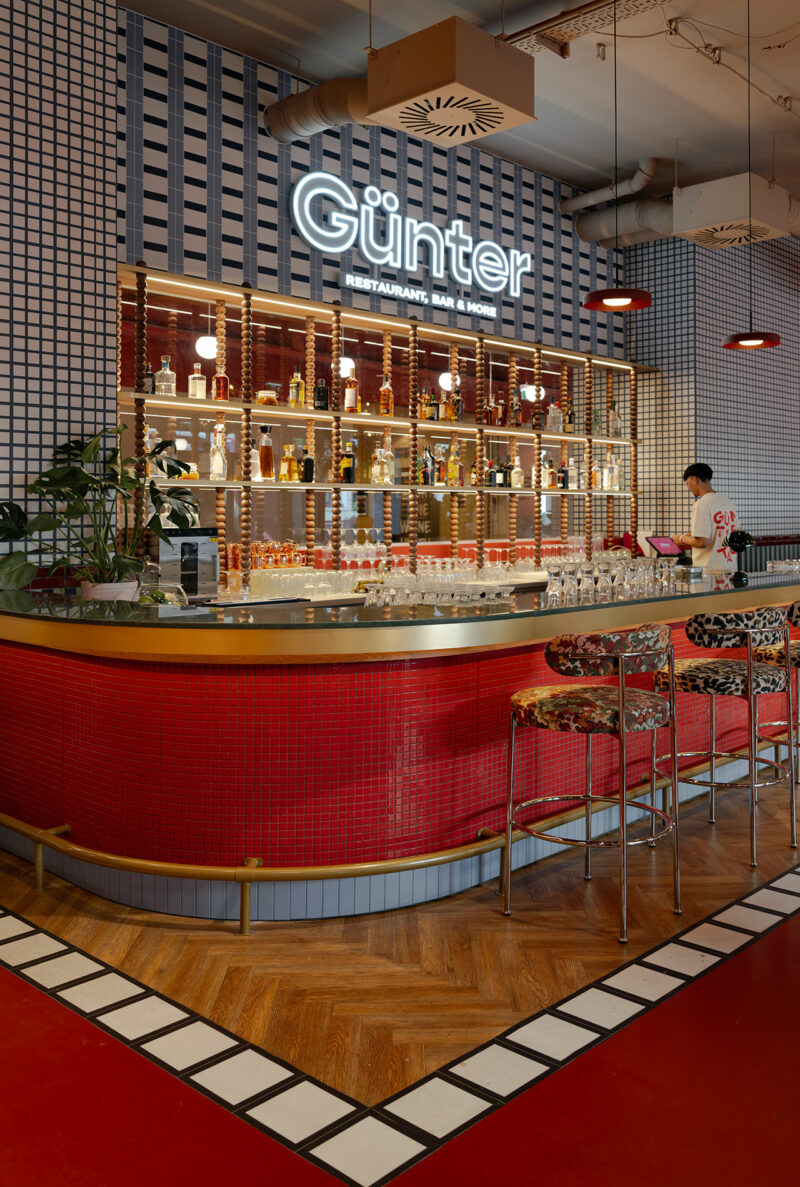 A bartender stands behind a red-tiled bar with a gold counter at Günter restaurant, designed by Atelier OLK. Shelves with bottles and a neon Günter sign glow in the background.