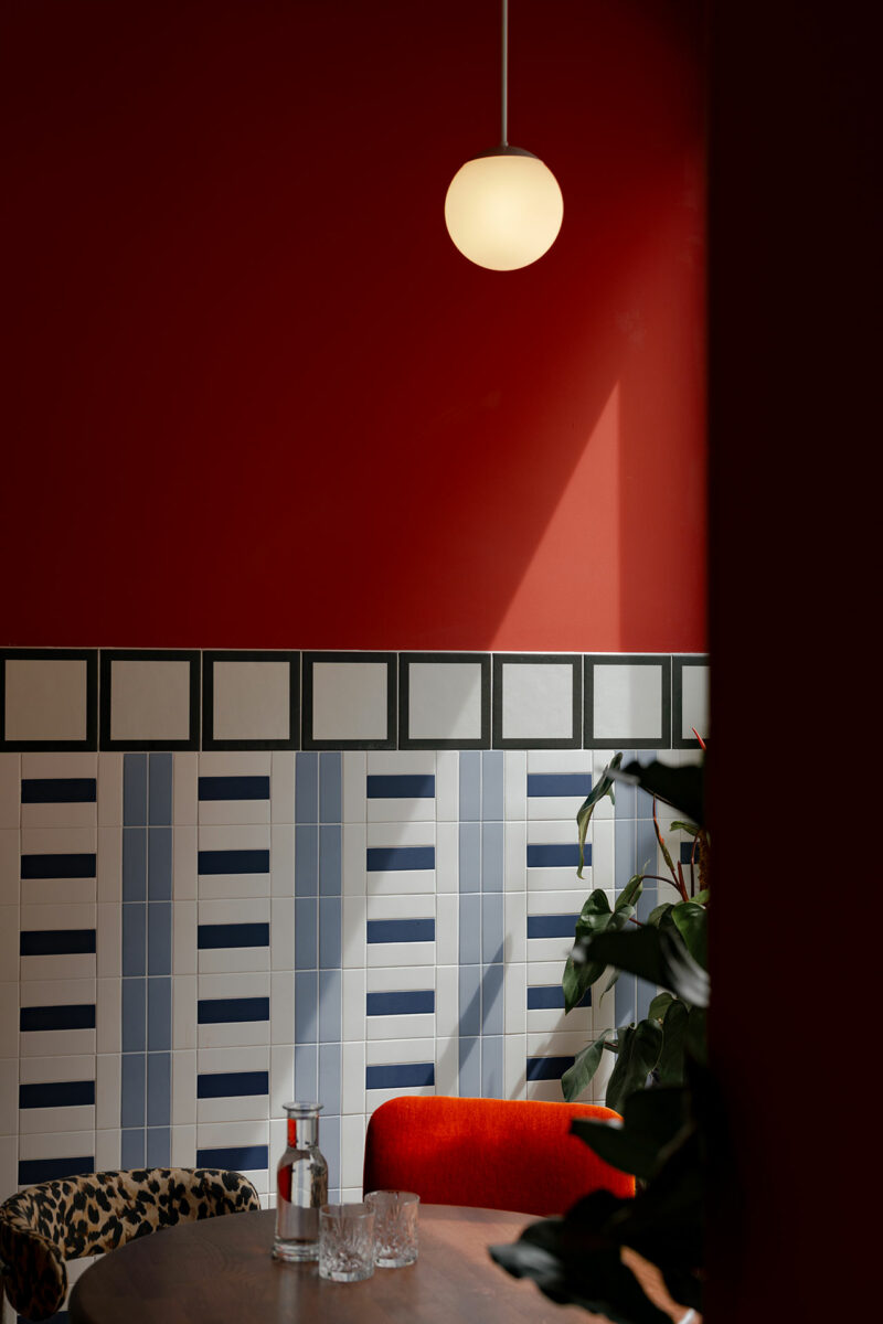 A round Atelier OLK pendant light hangs over a table with glassware, set against a red wall with geometric tile and a plant partially visible on the right.