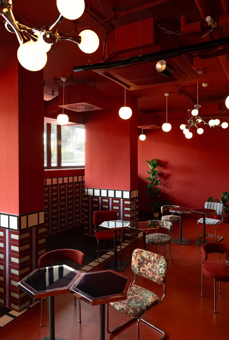 A modern café interior by Atelier OLK features red walls, geometric black and white tile accents, hexagonal tables, assorted chairs, and globe pendant lights.