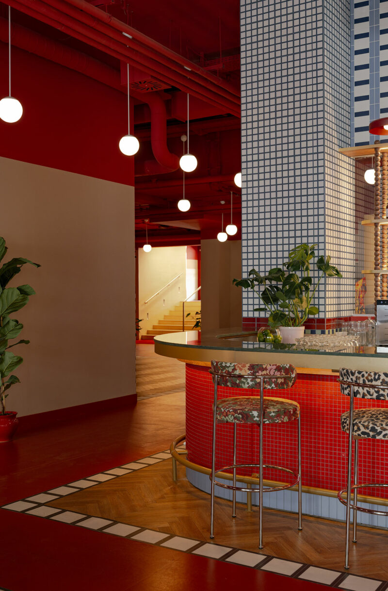 A modern bar by Atelier OLK features red tile, patterned barstools, round hanging lights, potted plants, and grid-patterned columns in a vibrant interior space.