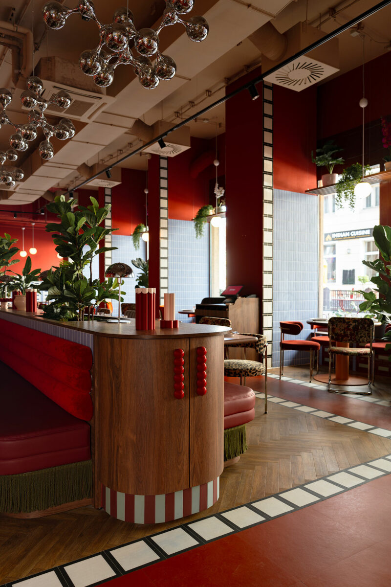 A modern restaurant interior by Atelier OLK with red and wood accents, globe light fixtures, green plants, and large windows letting in natural light.