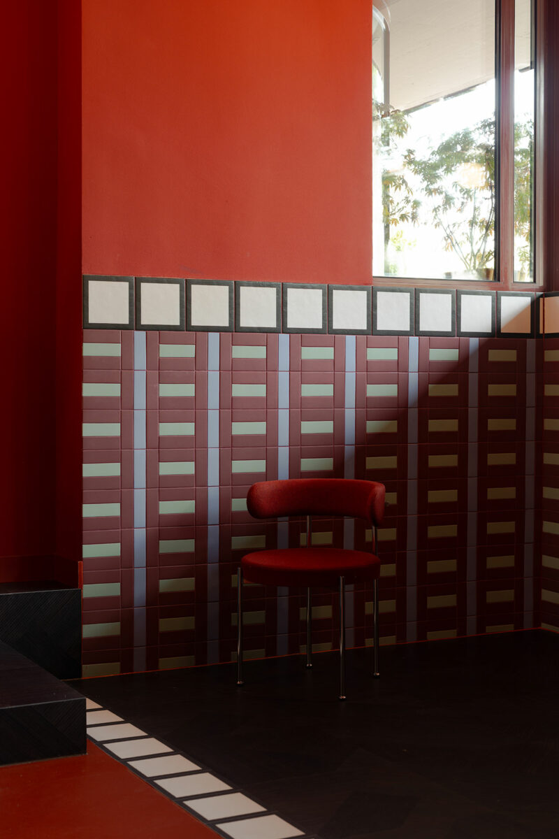 A red chair by Atelier OLK sits against a wall with geometric red and white tile patterns beneath a red-painted section, near a window with trees visible outside.