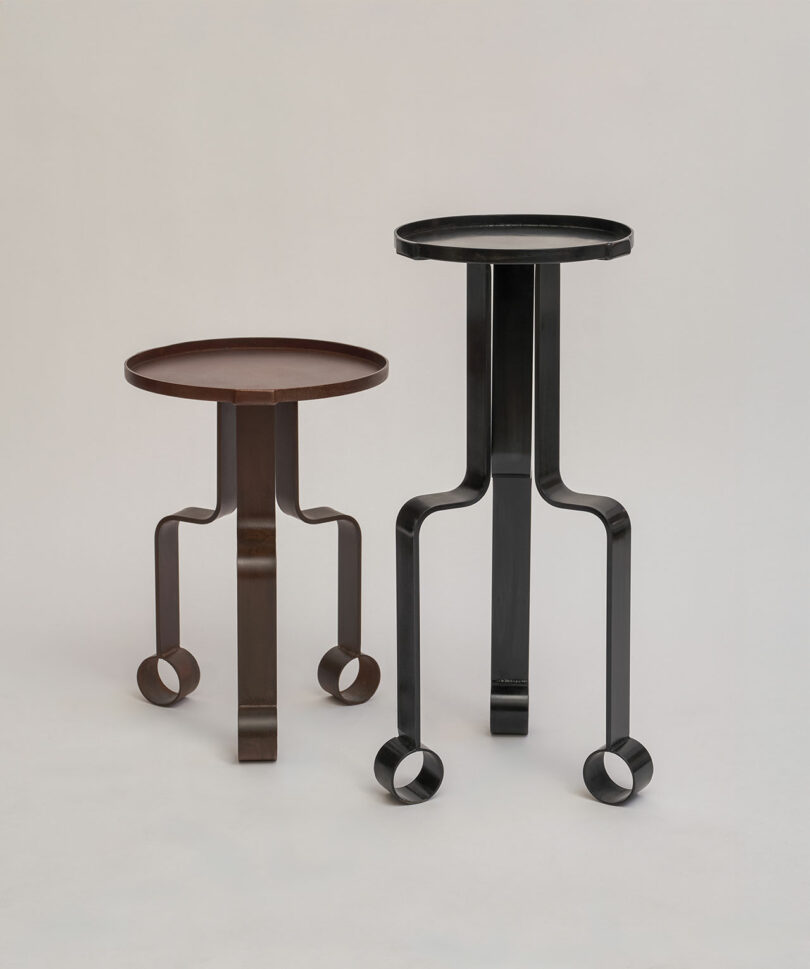Two metal side tables, one brown and one black, each with three curved legs ending in circular loops, stand on a plain, light-colored background.
