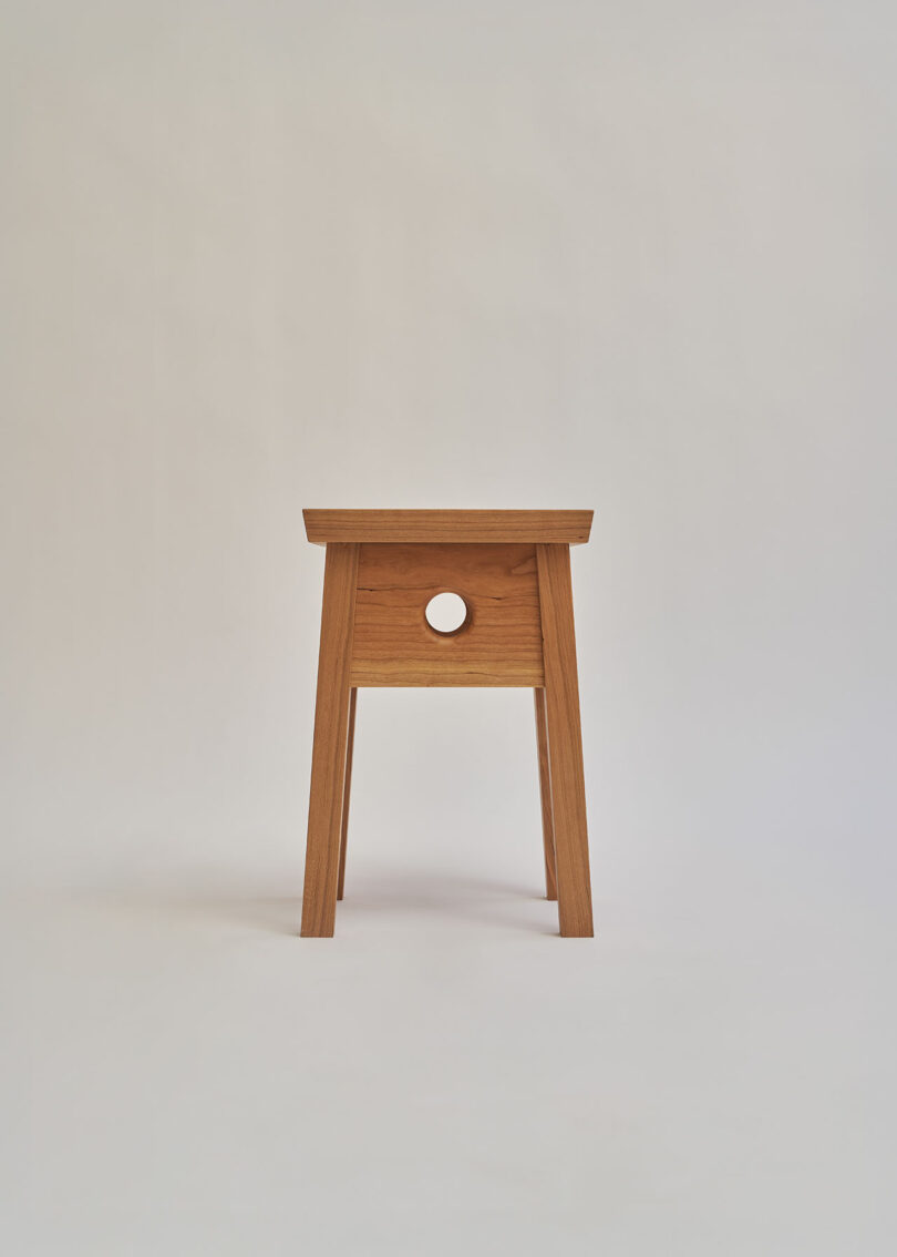A wooden stool with four legs and a rectangular seat, featuring a circular hole in the center of the front panel, is shown against a plain light background.
