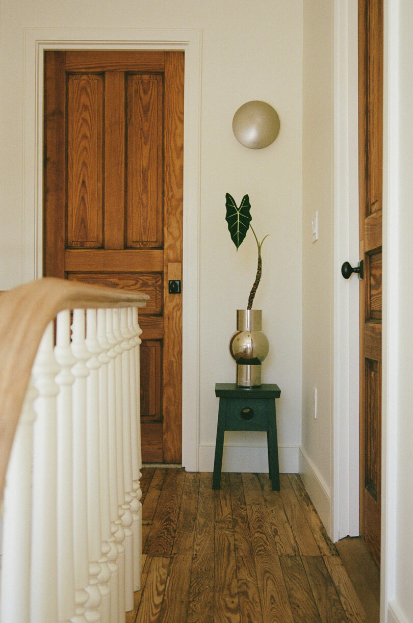 A small hallway with wooden floors and doors, a white railing, and a dark stool holding a potted plant under a round wall light.