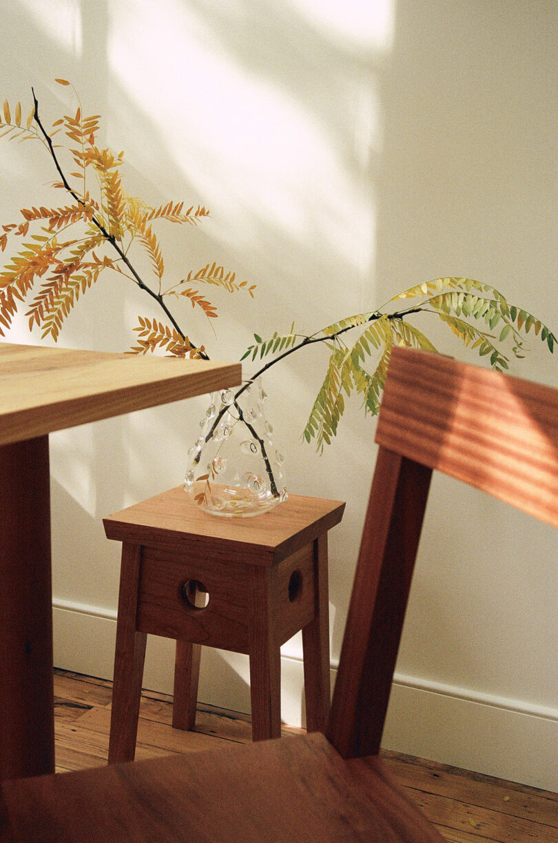 A wooden stool holds a clear glass vase with a leafy branch, next to a table and chair, sunlight casting shadows on the wall and furniture.