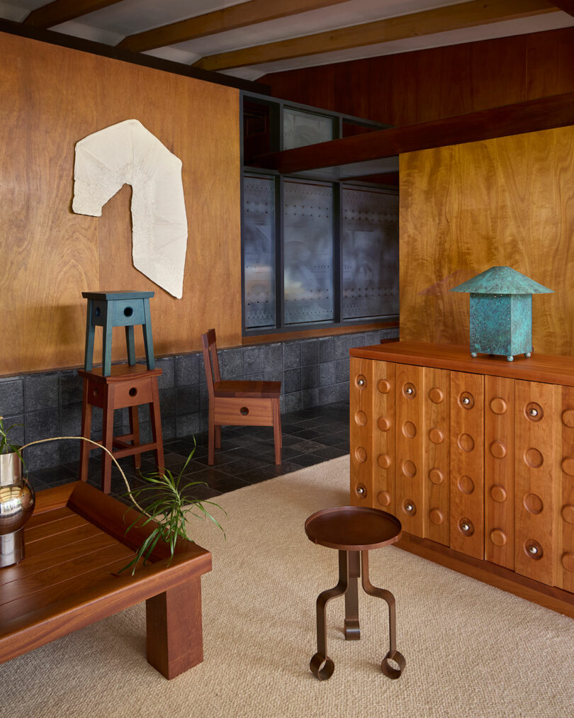 Mid-century modern interior with wood-paneled walls, geometric furniture, a cabinet with circular details, and a textured wall art piece above two stacked stools.