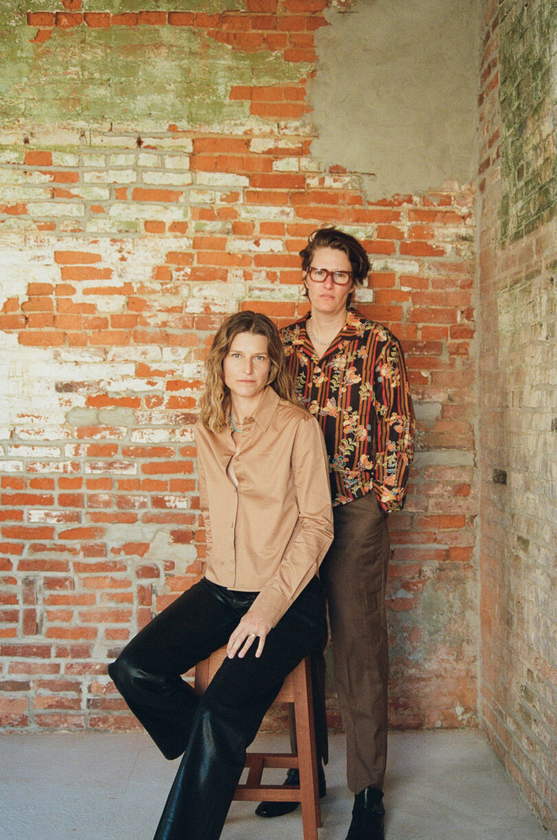 Two people pose indoors against an exposed brick wall; one is seated on a stool wearing a tan shirt, the other stands beside them in a patterned shirt and glasses.