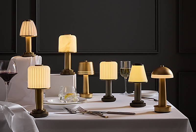 A variety of small, gold and brass table lamps with glowing shades arranged on a white tablecloth, set with plates, glasses, and cutlery against a dark wall.