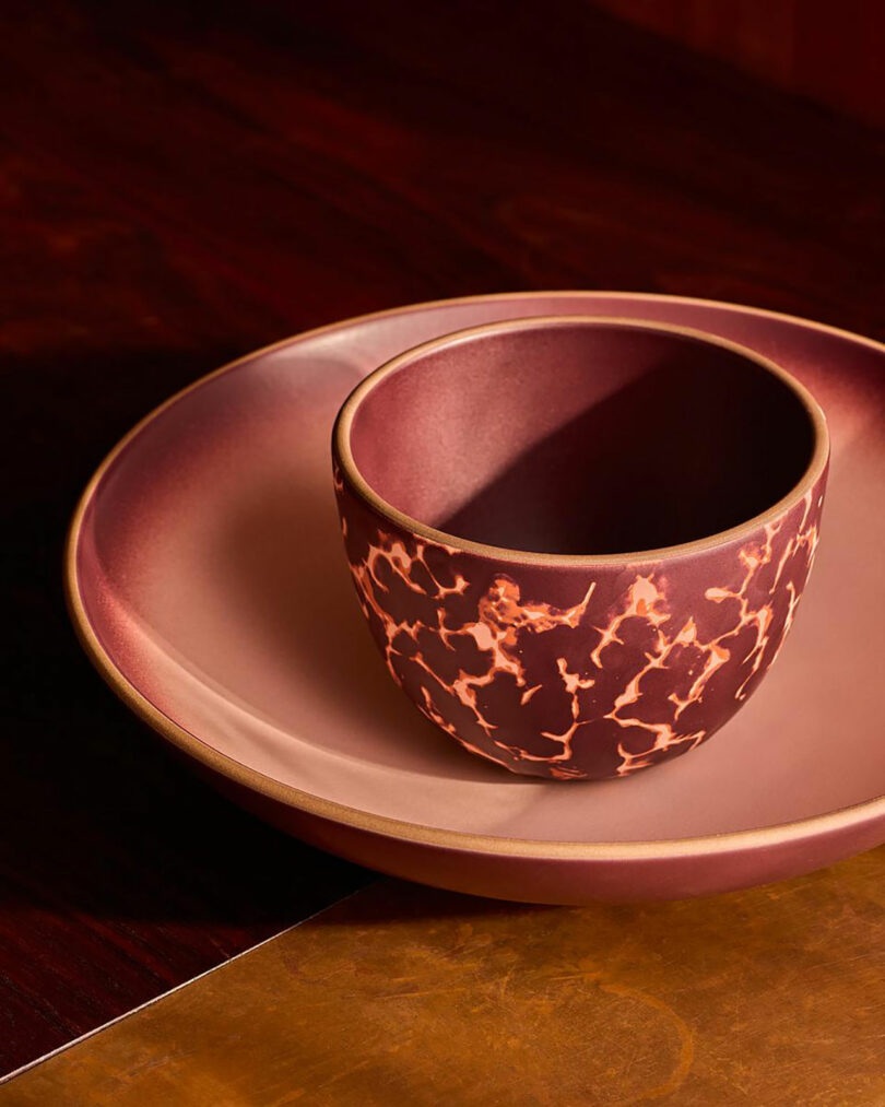 A maroon ceramic cup with a marbled design sits inside a matching maroon plate on a wooden surface.