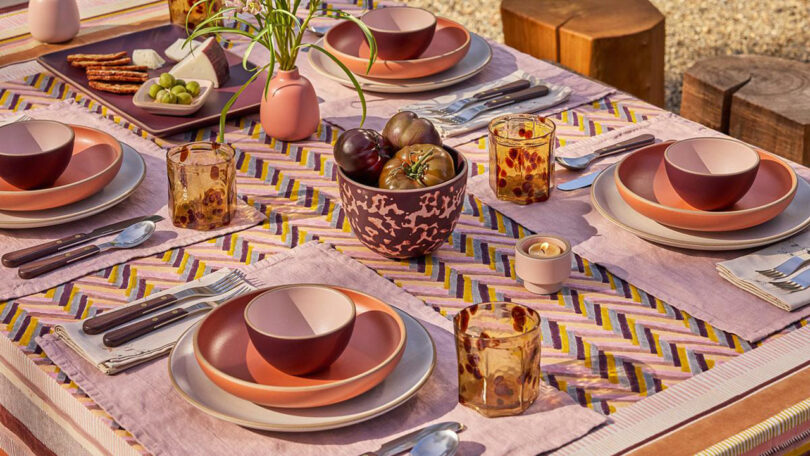 A table set outdoors with patterned cloth, plates, bowls, glasses, cutlery, a vase with flowers, a bowl of fruit, and small food platters arranged neatly for four people.