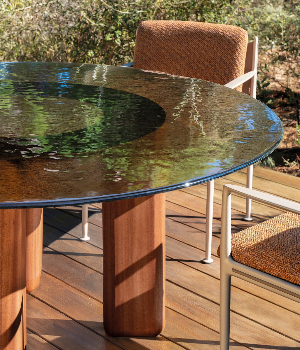 A round glass table with wooden legs and cushioned chairs sits on a wooden deck, reflecting trees and greenery in its surface.