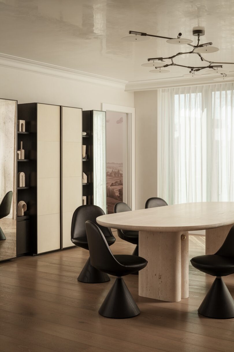 A modern dining room with a large oval table, six black chairs, tall shelving units, floor-to-ceiling curtains, and a geometric ceiling light fixture.