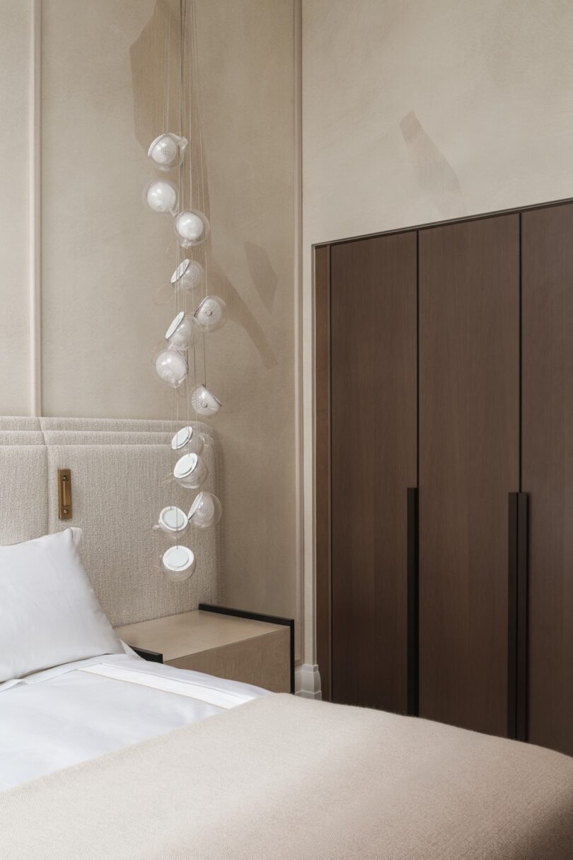 A modern bedroom with a beige bed, a unique hanging glass pendant light, and a tall wooden wardrobe against neutral walls.