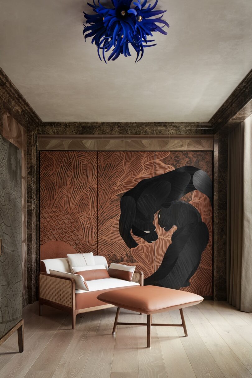 Modern lounge area with a tan armchair and ottoman, patterned wood wall featuring black abstract art, light wood flooring, and a blue sculptural ceiling light.