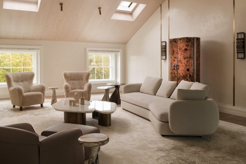 Bright, modern living room with a beige sofa, two textured armchairs, a marble coffee table, and skylights on a sloped wooden ceiling. A decorative cabinet stands by the wall.