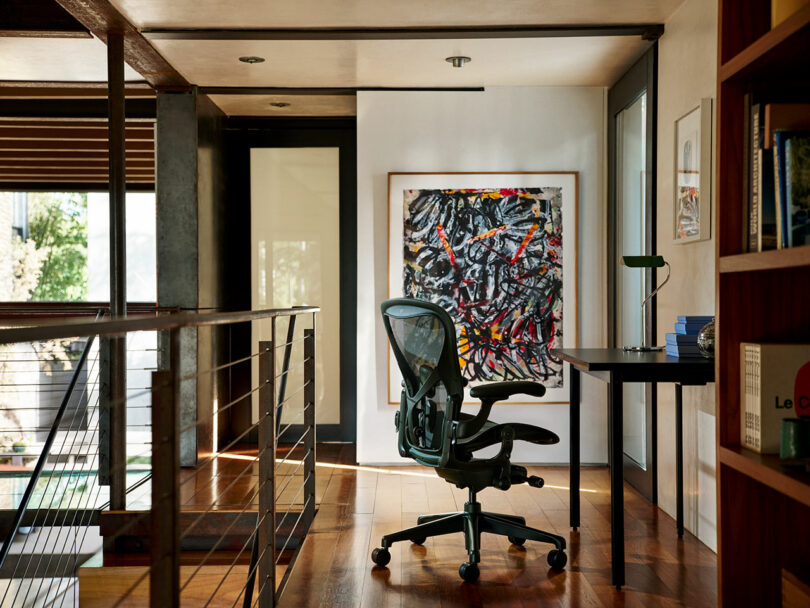 A modern home office with a black ergonomic chair, a black desk with a lamp and books, wooden floors, and an abstract painting on the wall.