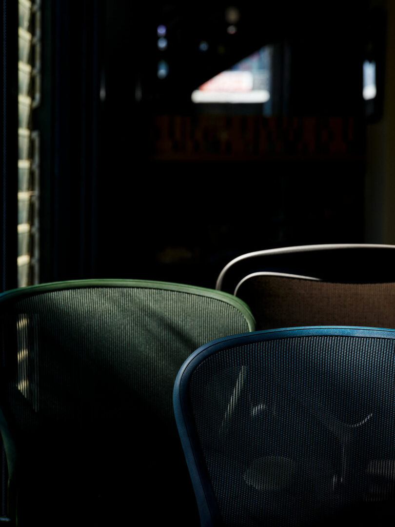 Three empty mesh office chairs in green, brown, and blue are partially illuminated by light, set against a dark background.
