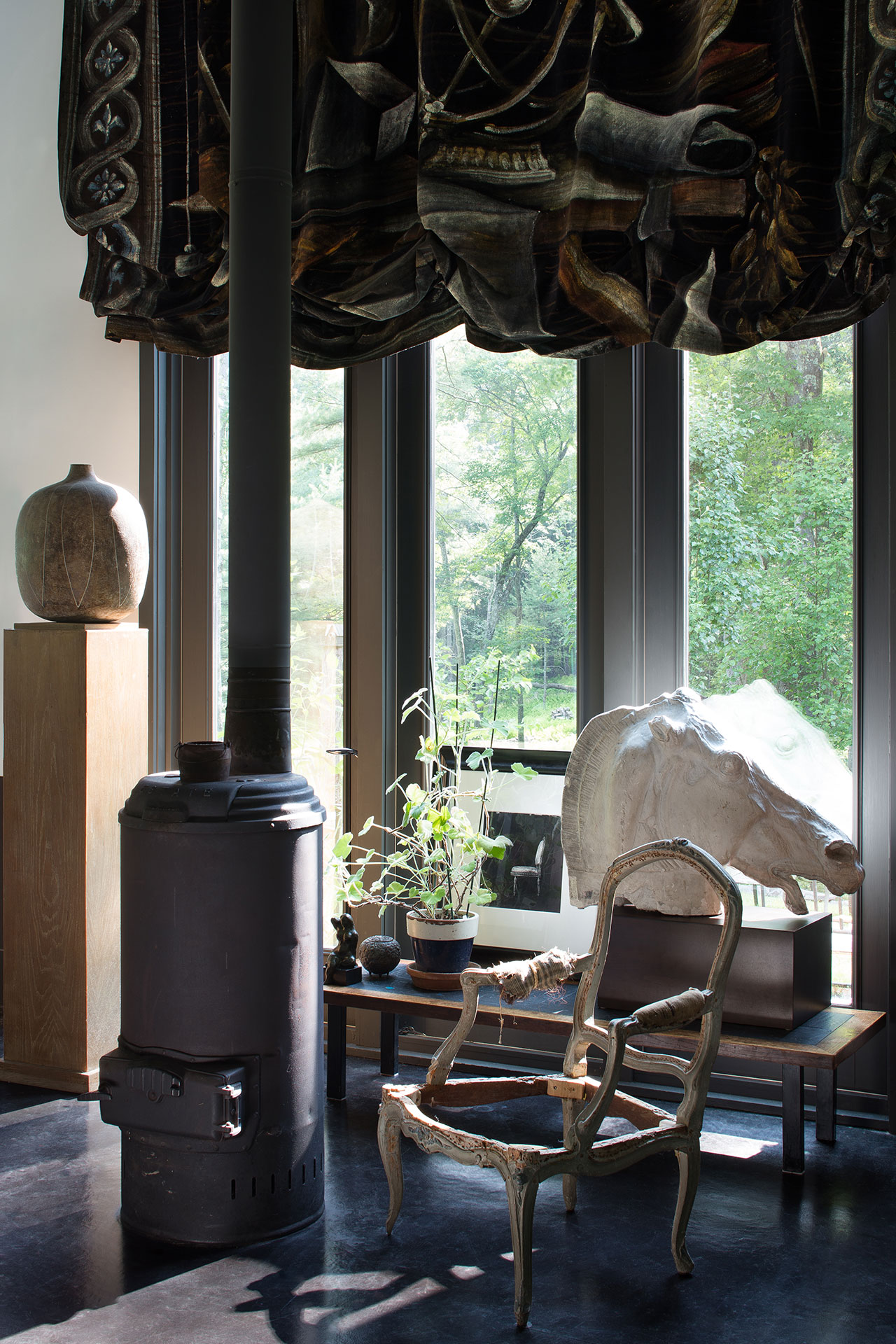A sunlit corner with large windows features a wood stove, a partially stripped antique chair, potted plants, a large animal skull, a ceramic vase, and a decorative curtain.
