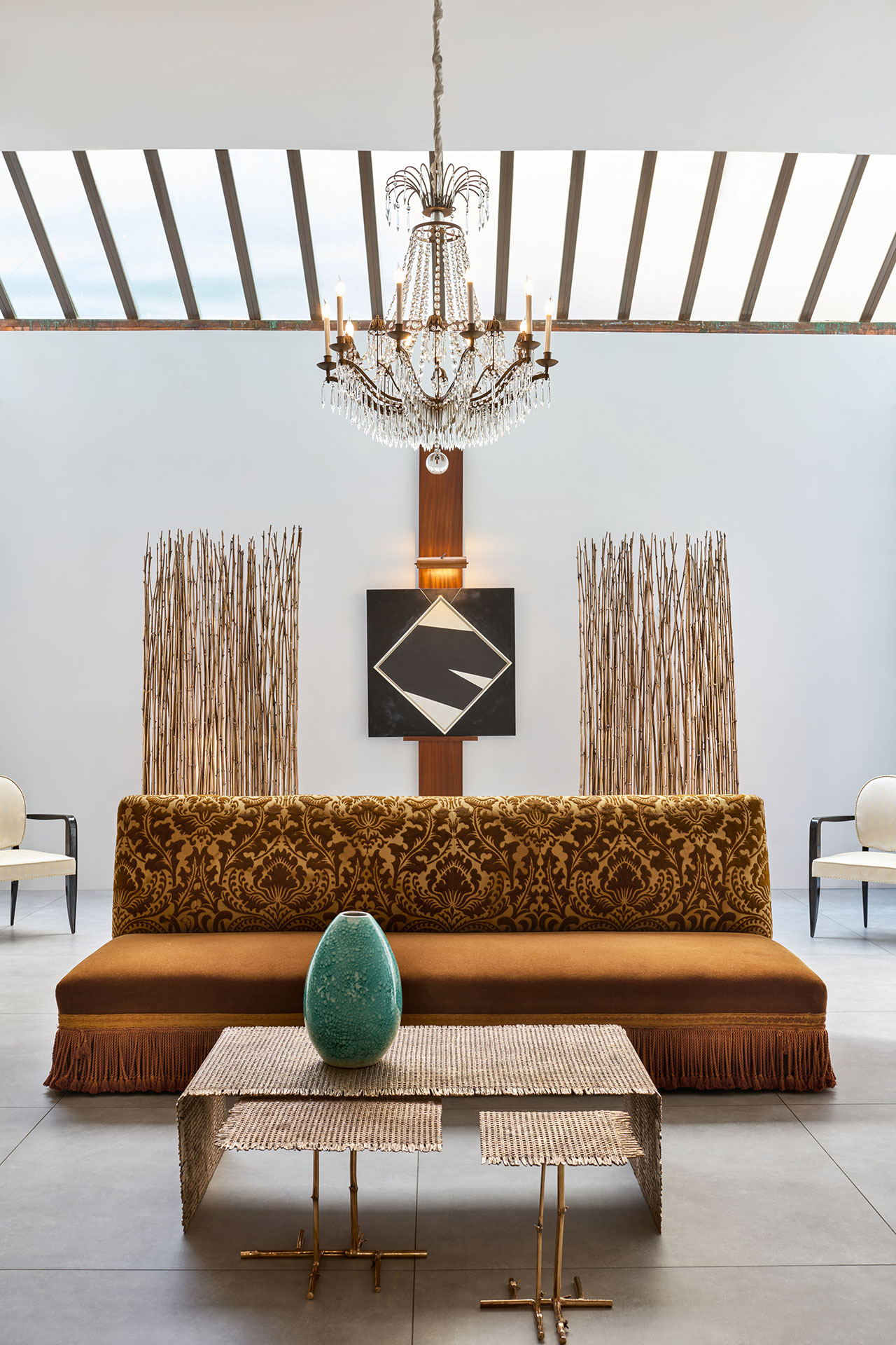 A modern living room with a gold patterned sofa, glass chandelier, geometric wall art, two white chairs, a turquoise vase on metal tables, and vertical wooden accents.