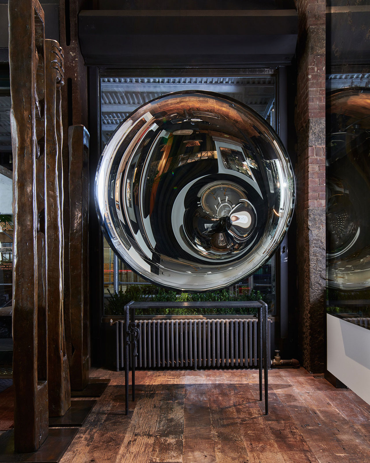 A large, reflective, spherical sculpture is displayed indoors above a radiator, with exposed brick walls, wooden flooring, and large windows in the background.