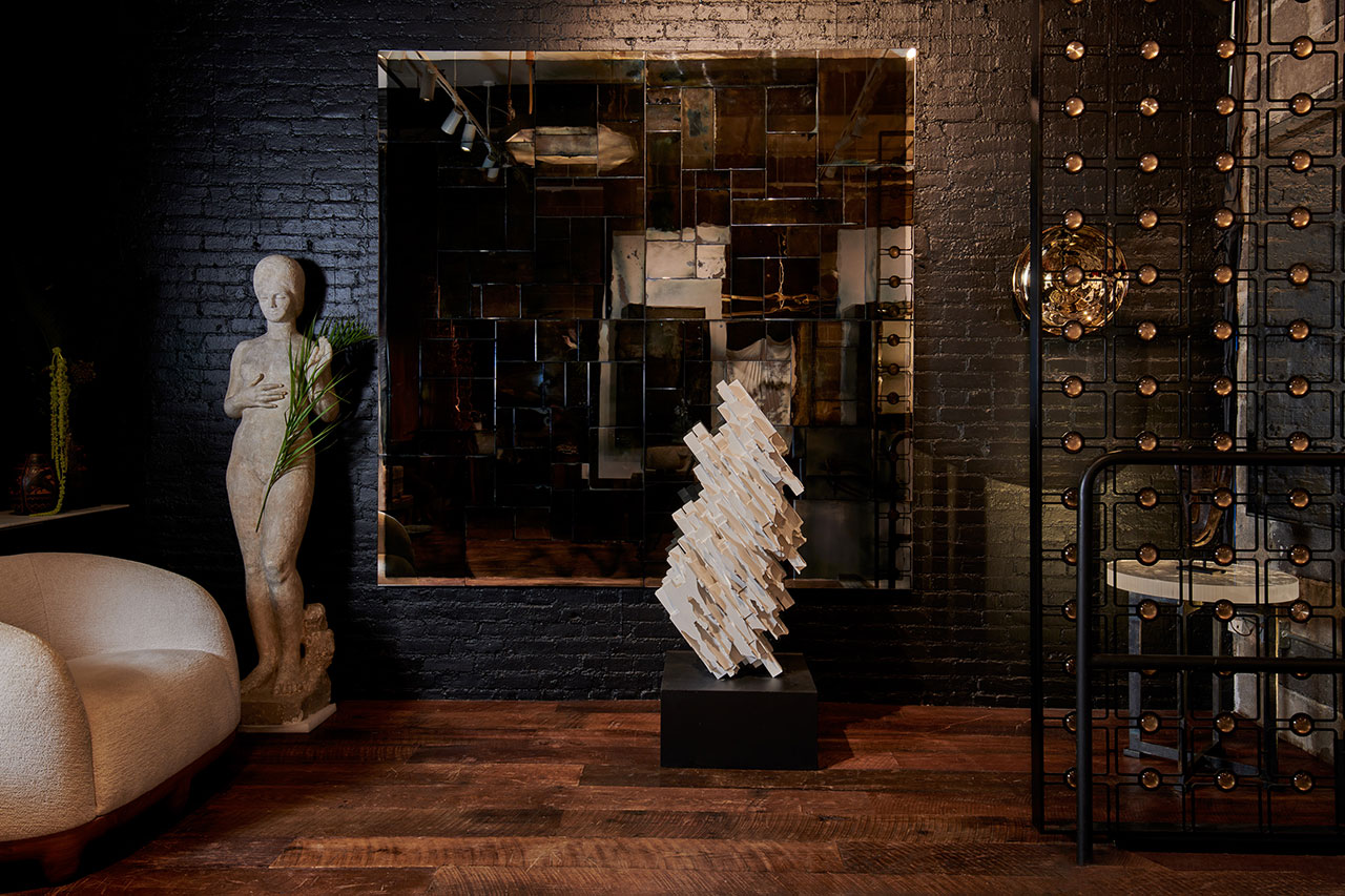 Modern interior with a white statue, a large geometric mirror, a sculptural piece on a pedestal, and a white armchair against a dark brick wall and wooden floor.