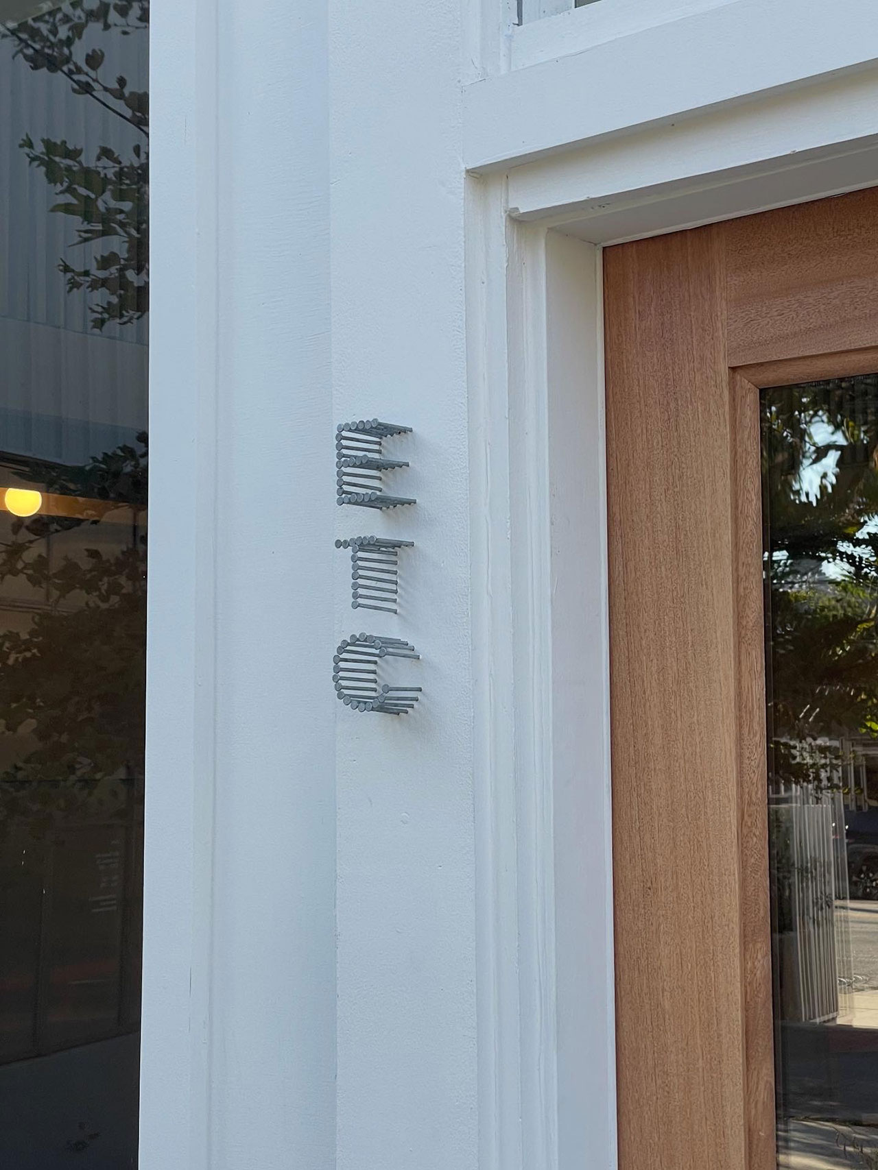 Vertical sign on a white wall next to a wooden door, with the word "ETC" spelled out in large silver nailsâeach one like a building block creating a unique statement piece.