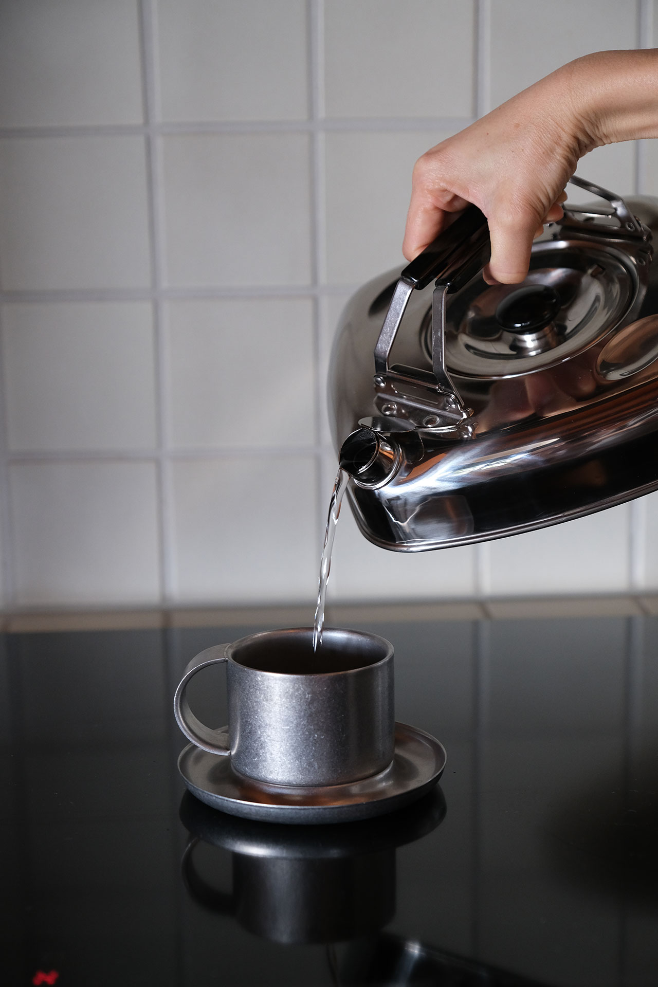 A hand pours hot water from a metal kettle into a gray cup on a saucer, with a tiled wall in the backgroundâa simple building block of daily rituals.