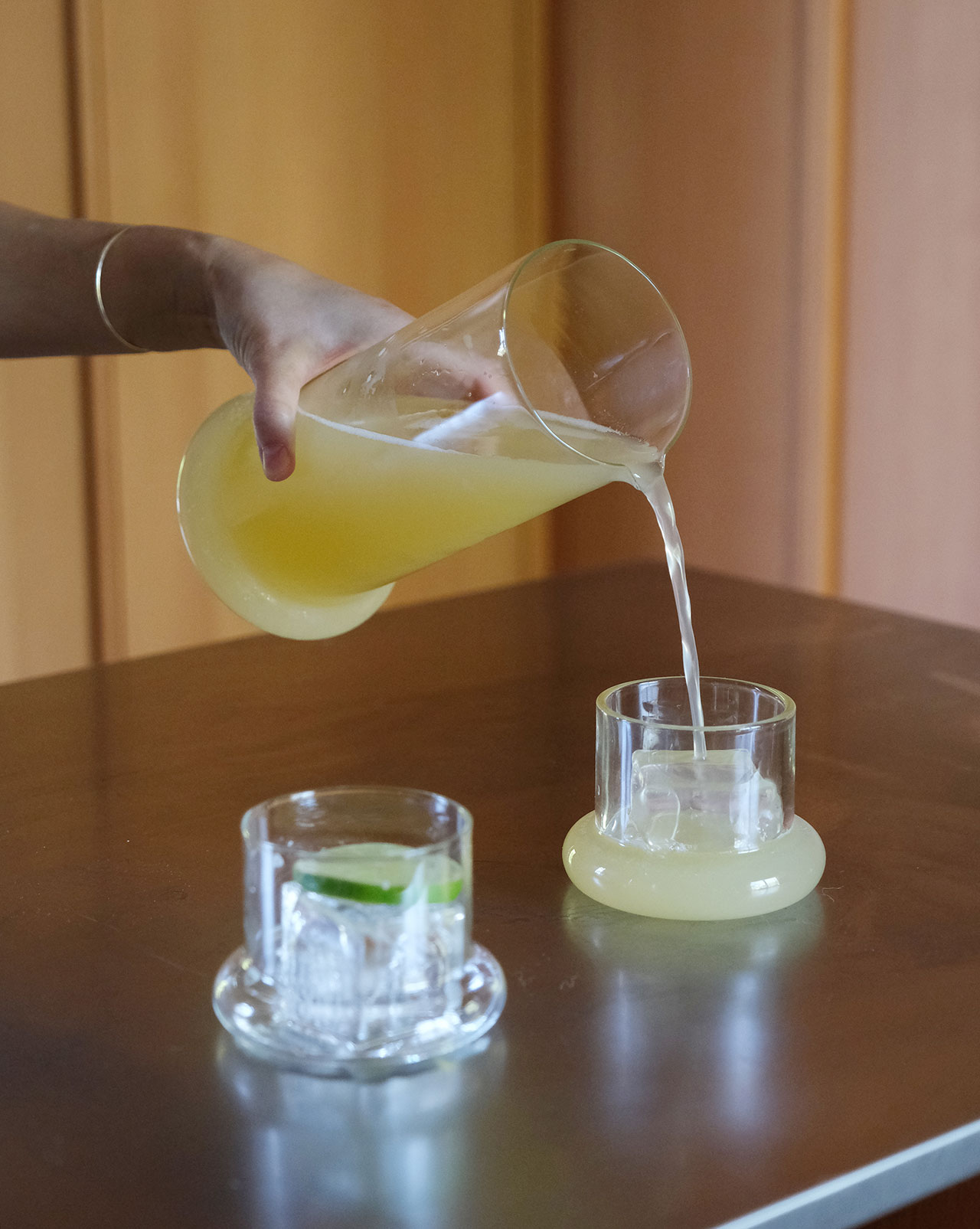 A hand pours yellow juice from a glass pitcher into a glass on a wooden tableâa building block of any summer gathering; another glass with ice and a lime wedge sits beside it.
