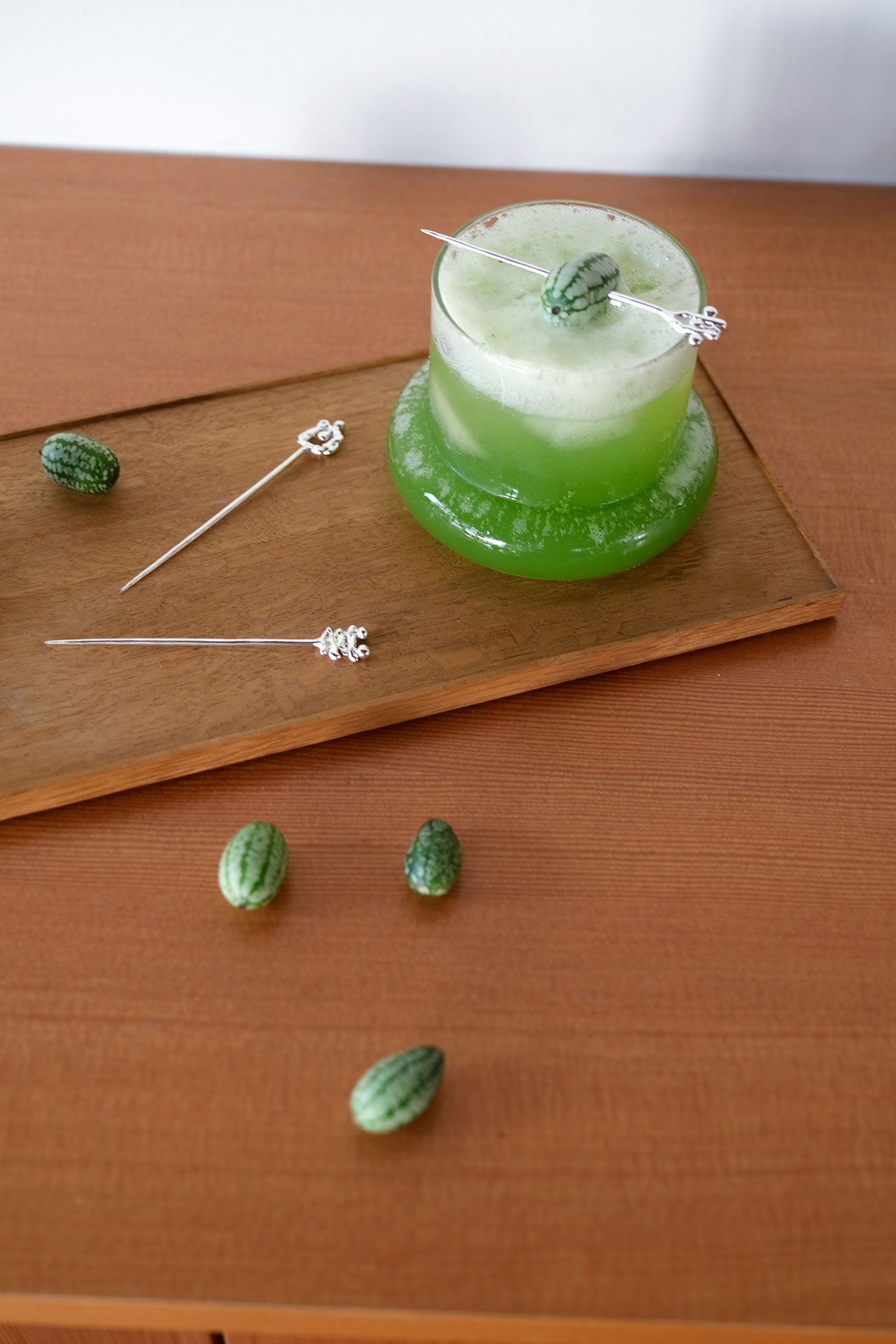 A green cocktail with a frothy top sits on a wooden tray, garnished with a mini cucumber on a cocktail pickâeach ingredient serving as a building block of fresh flavor. Three more mini cucumbers and two picks are on the tray and table.