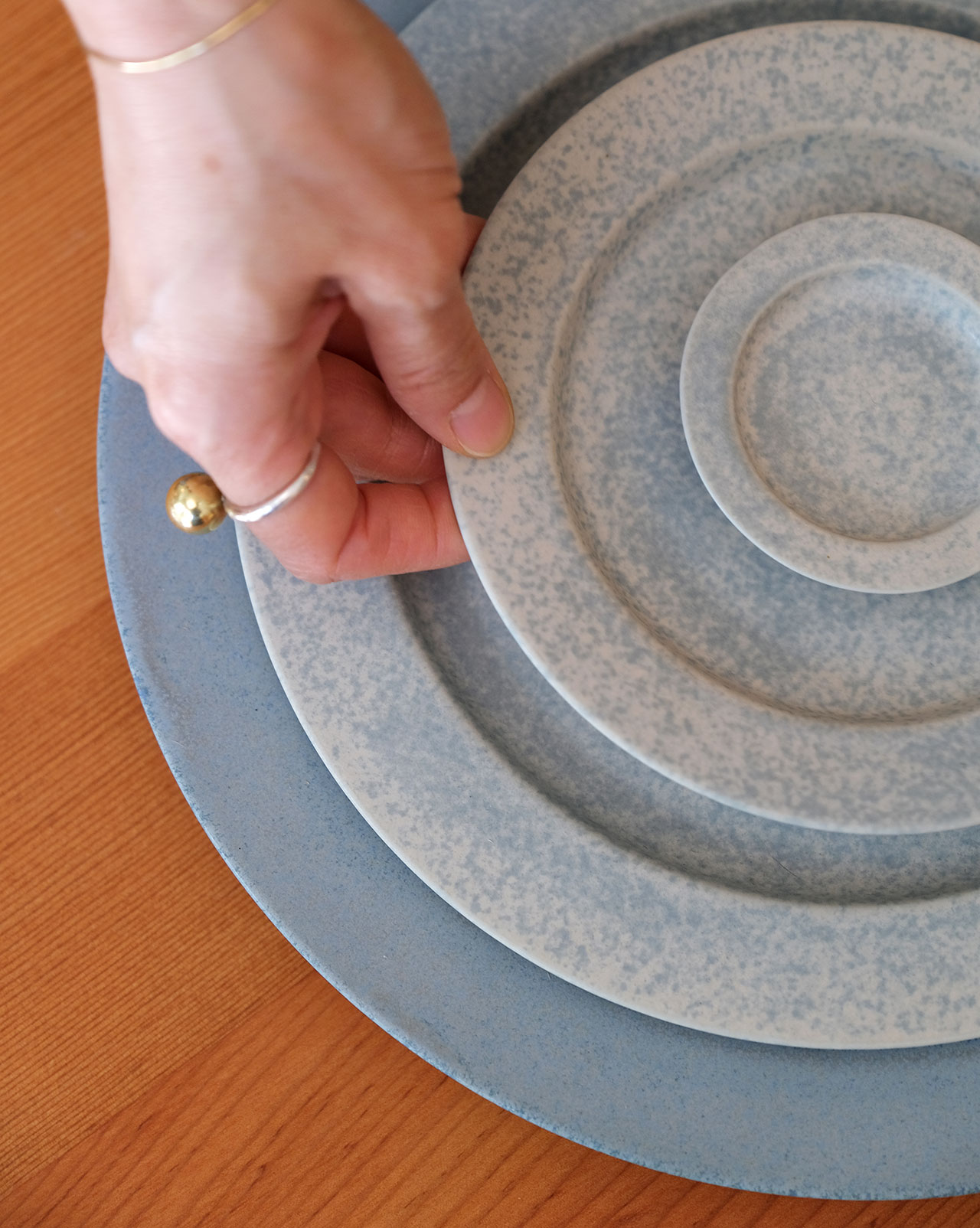 A hand holding a small speckled plate, stacking it on larger plates arranged in descending size order on a wooden surfaceâlike building blocks for a beautiful table setting.