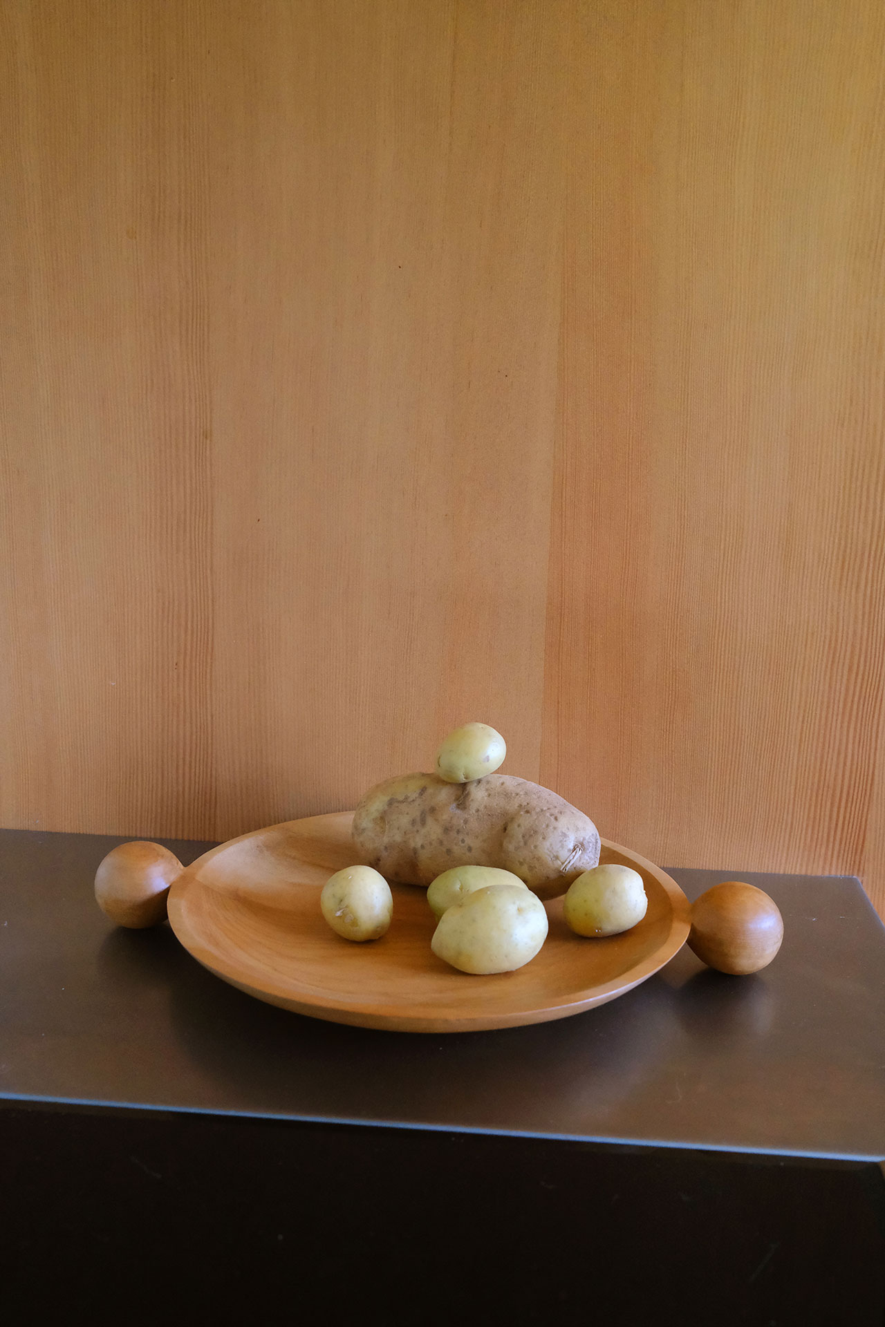 A wooden plate with six potatoes of varying sizes, each a humble building block of countless meals, sits on a dark surface against a light wood-paneled background.