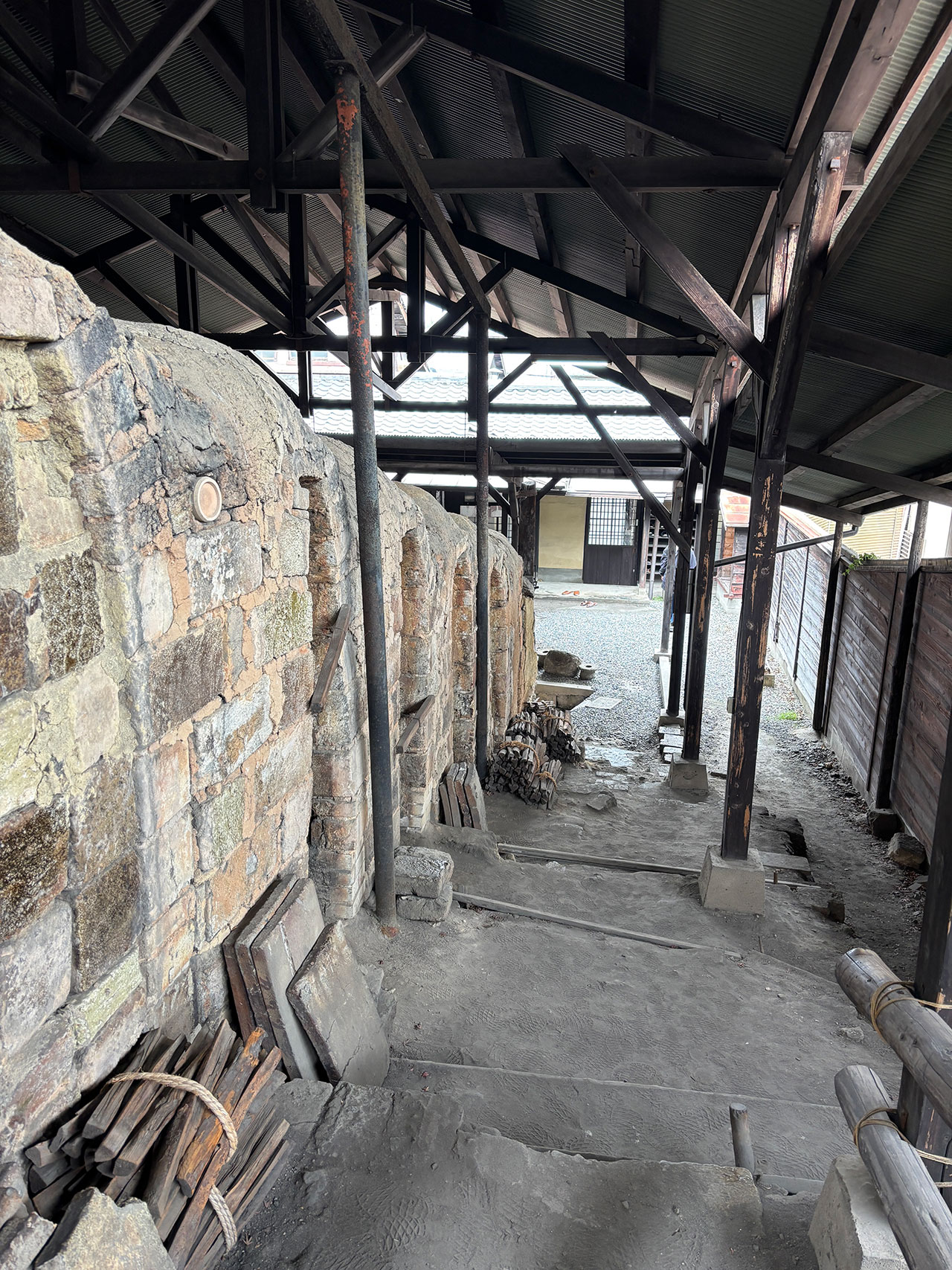 Rows of old stone kilns with wooden supports and scattered tools, sheltered under a metal roof in an outdoor workspace, evoke the traditional artistry of Kawai Kanjira.
