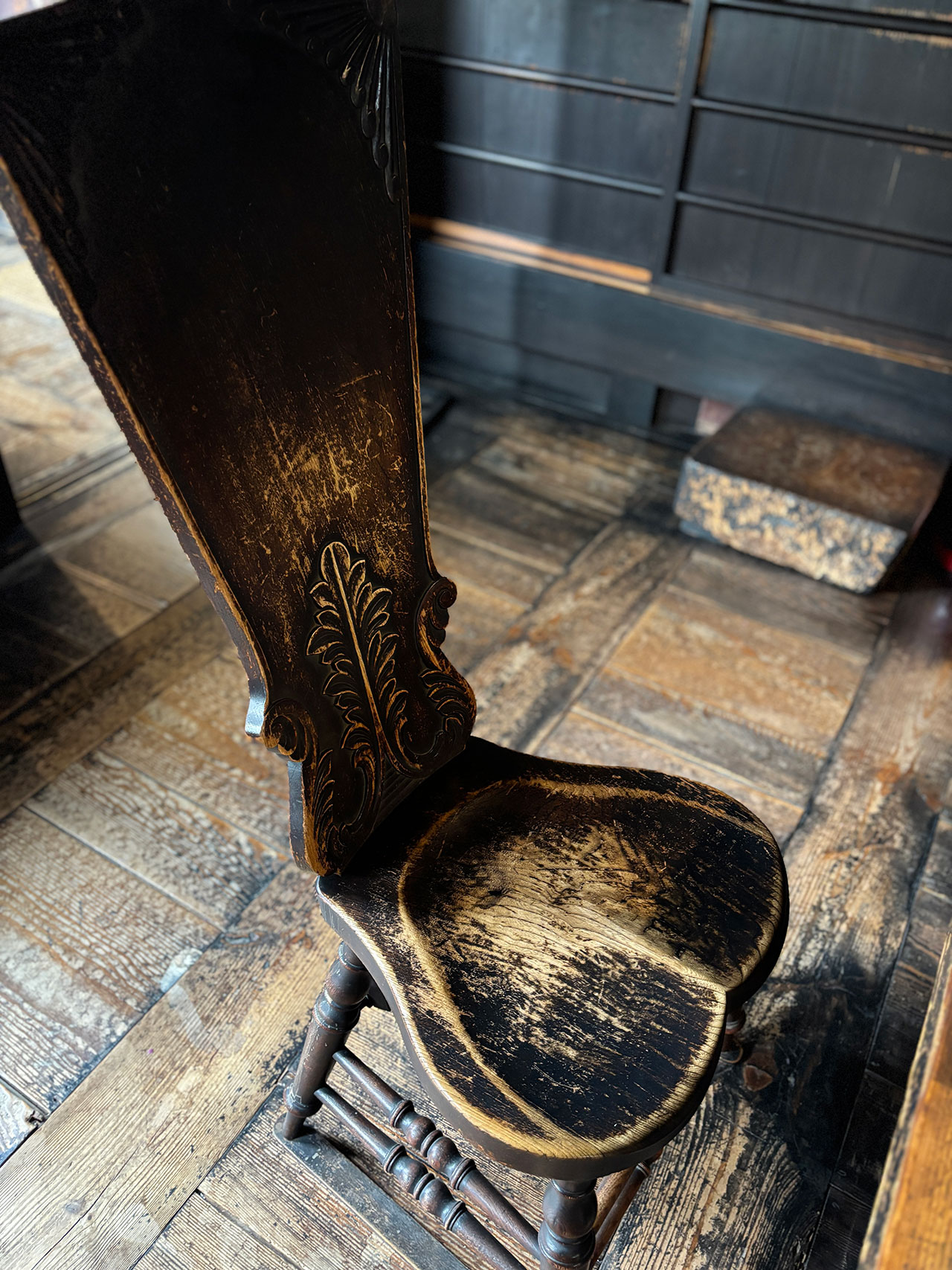 A worn wooden chair with ornate carvings on the backrest and a curved seat sits beside a Kawai Kanjira, both resting on a rustic wooden floor in a dimly lit room.