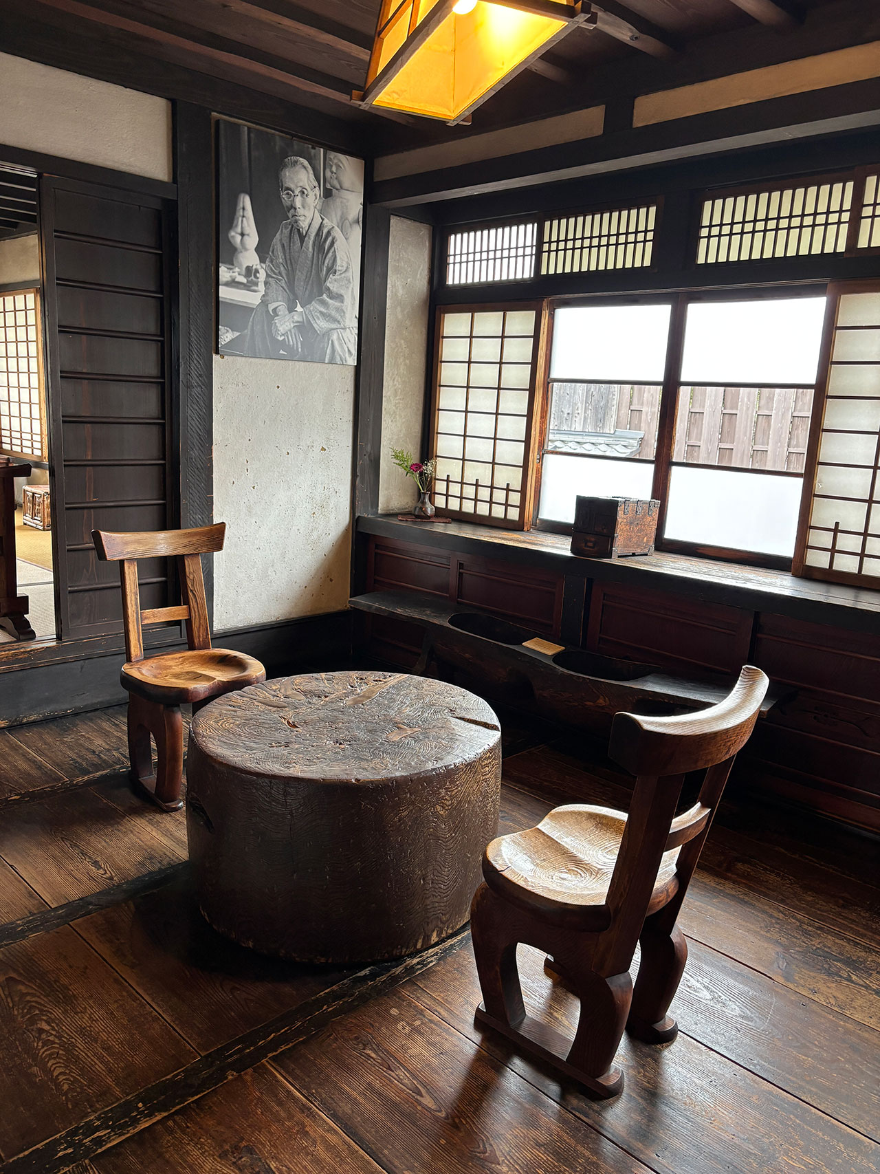 A wooden room with two chairs and a round table, large windows with wooden frames, a Kawai Kanjira resting nearby, and a black-and-white portrait of a man on the wall.