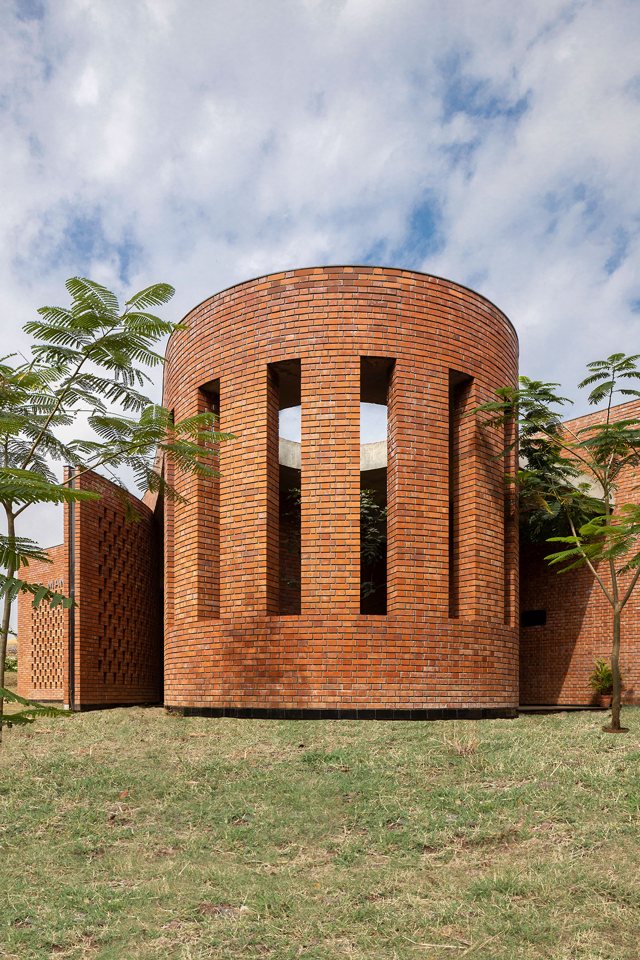 A cylindrical brick building, shaped by morphogenesis, features tall vertical windows and stands on a grassy area, with small trees nearby and a partly cloudy sky above.