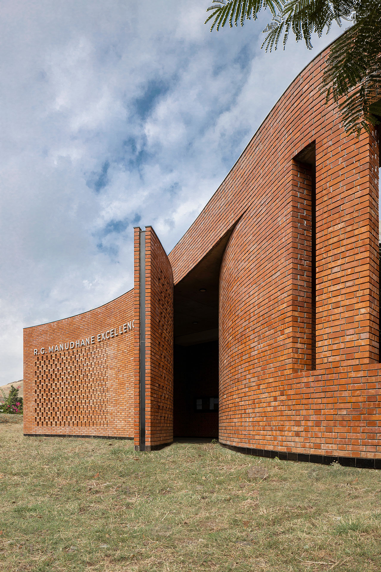 Curved brick building by Morphogenesis with perforated brickwork, labeled "R.G. MANUPHANE EXCELLENCE," set against a partly cloudy sky and grassy foreground.