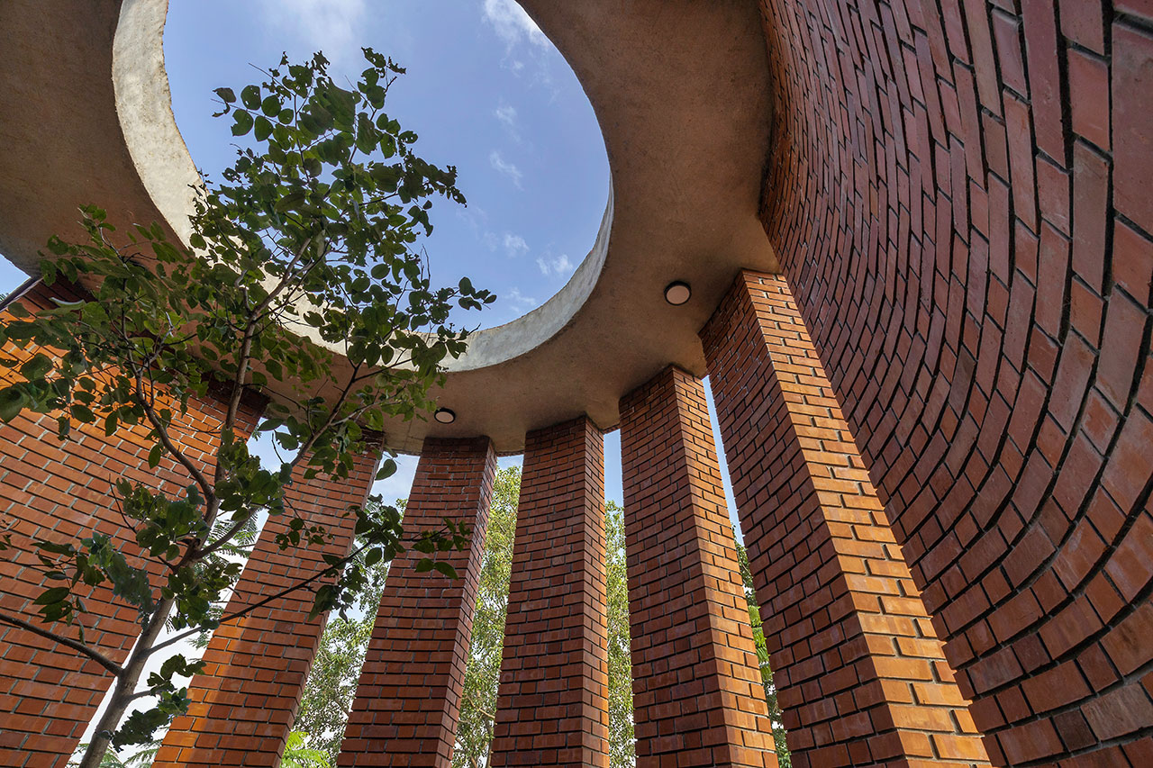 View from inside a circular brick structure with vertical pillars, an open roof featuring a large morphogenesis-inspired round cutout, and a tree growing inside against a blue sky.