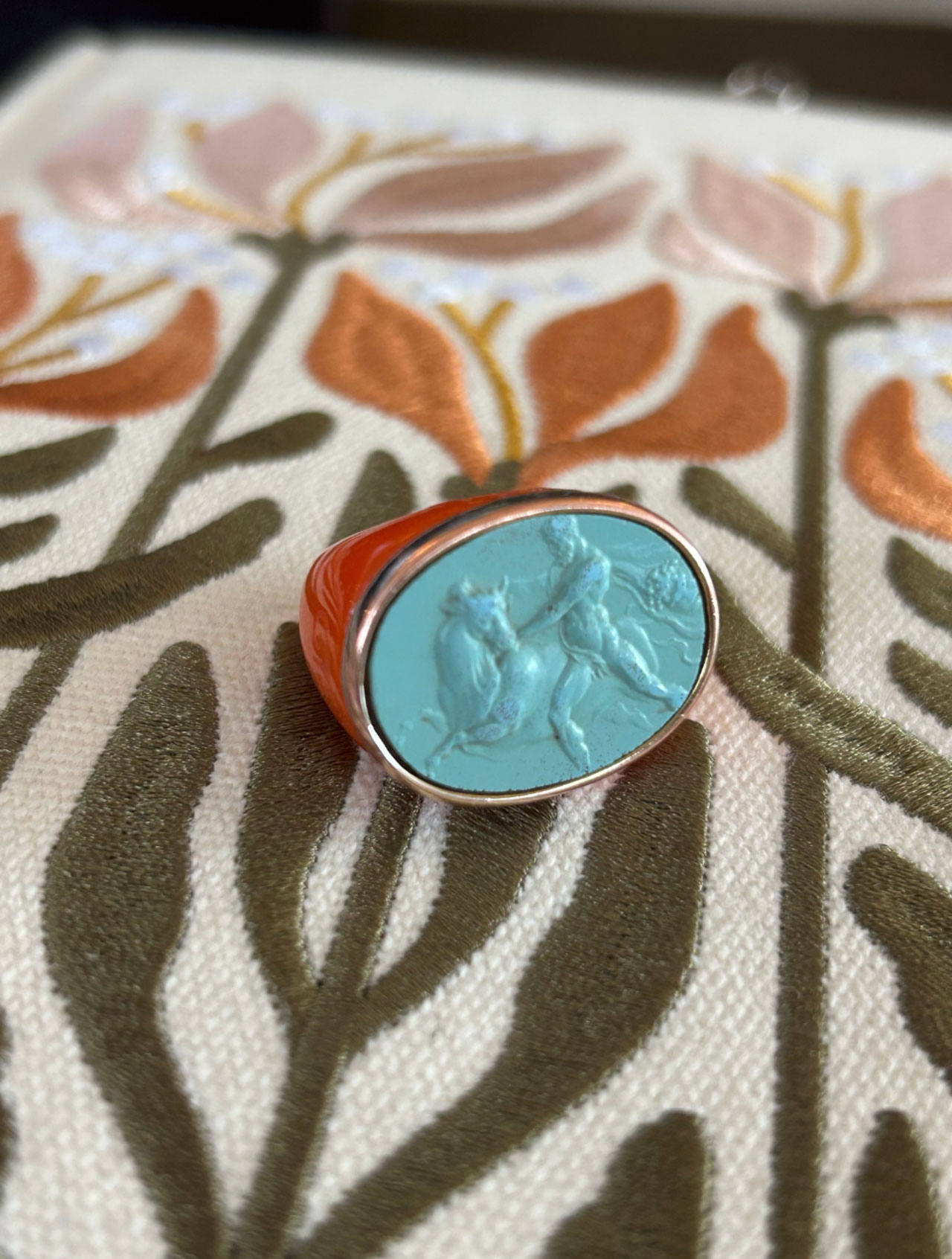 A signet ring with a light blue cameo depicting two figures is placed on an embroidered fabric with floral and leaf patterns in orange, brown, and green.