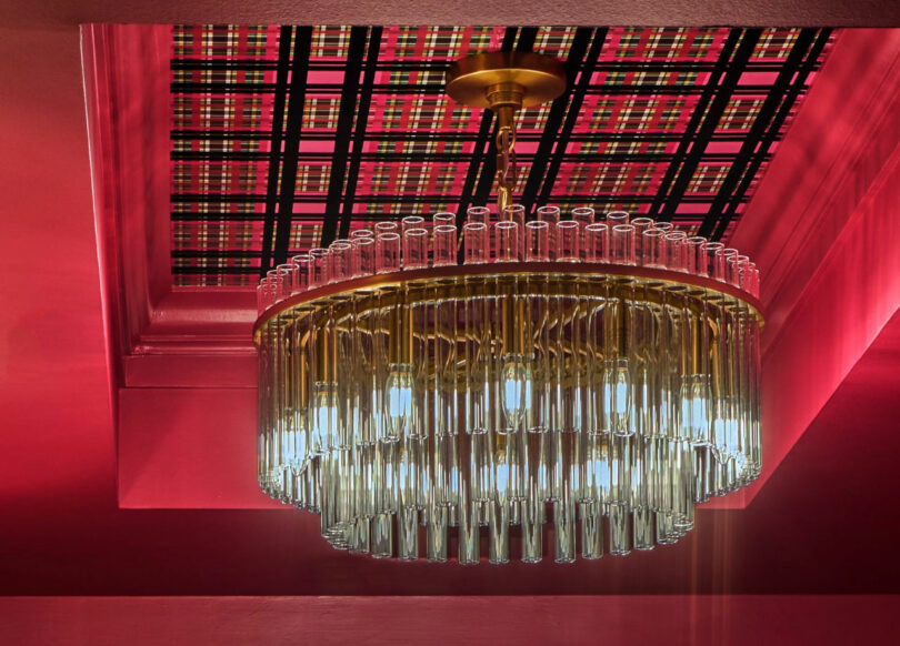 A modern chandelier with glass tubes hangs from a gold fixture, set against a red ceiling with a black and red plaid pattern inset.