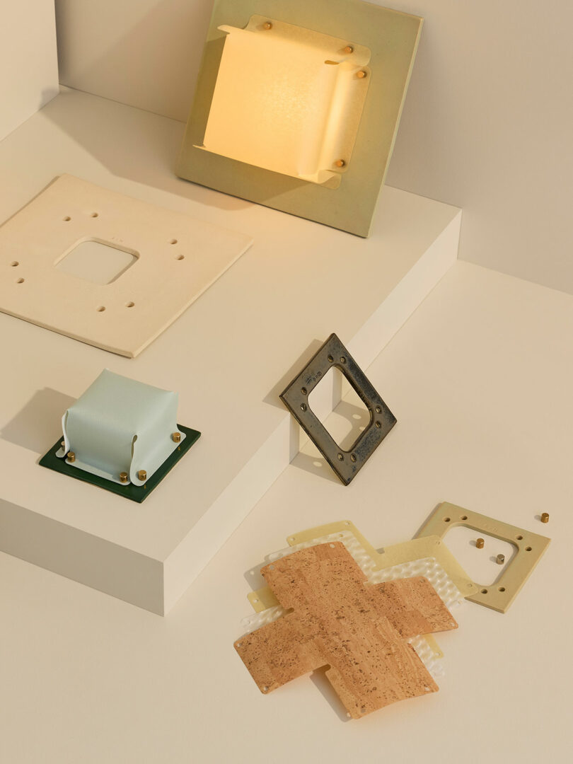 Geometric objects made of materials like cork, plastic, and metal are arranged on a cream-colored tiered surface with soft lighting.