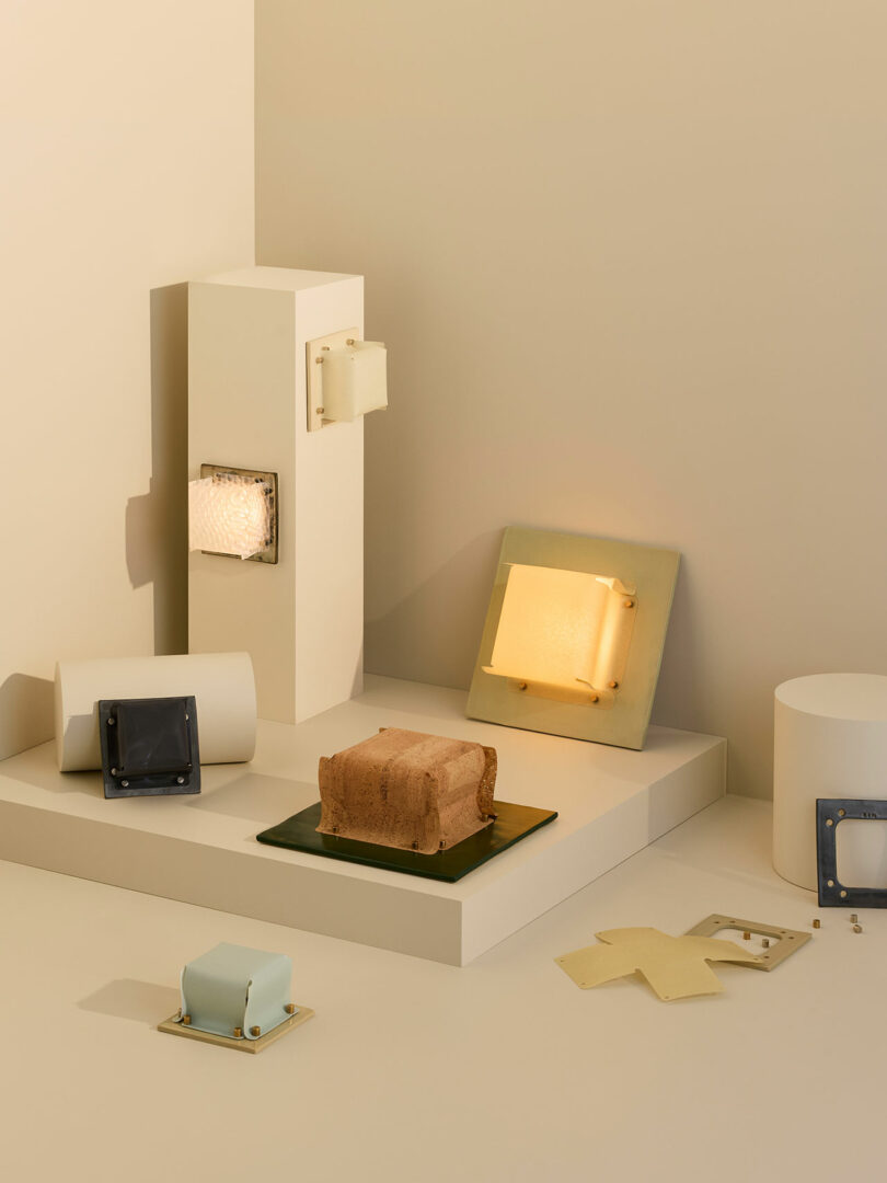 A minimalist display of geometric objects and wall-mounted lamps in neutral tones, with a clay block and various square and rectangular fixtures arranged on white platforms.